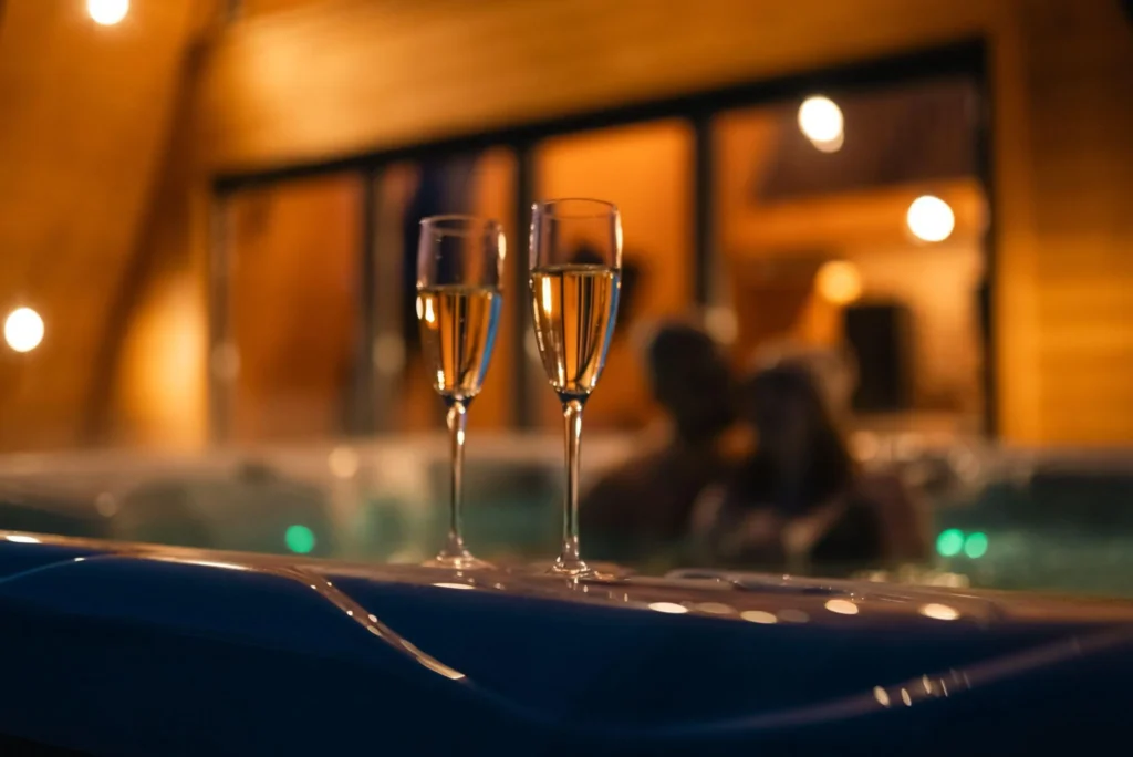 Two glasses of champagne on the edge of a hot tub at dusk, with a couple relaxing in the warm-lit water behind