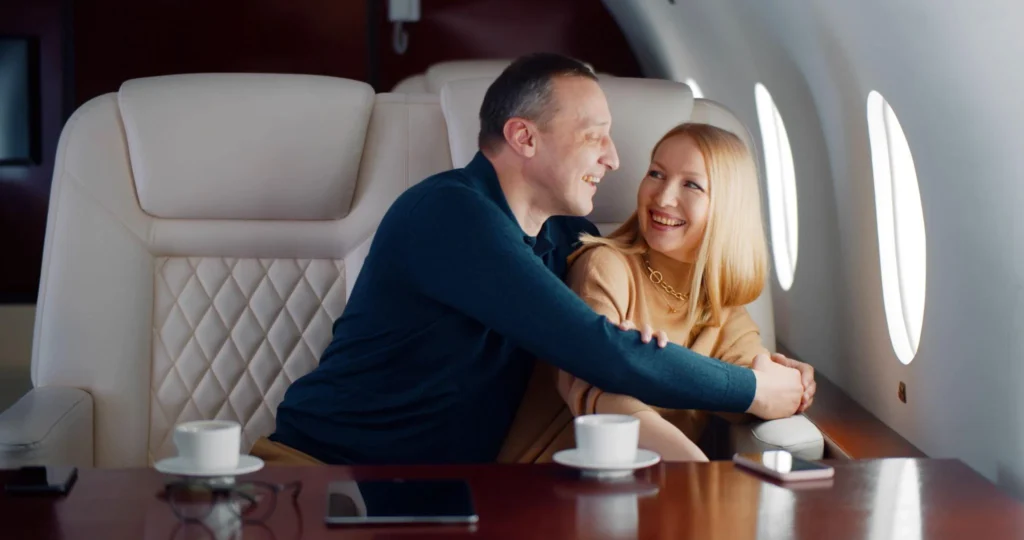 Smiling couple embracing in the cream leather seats of a private jet cabin, with coffee cups on the polished table