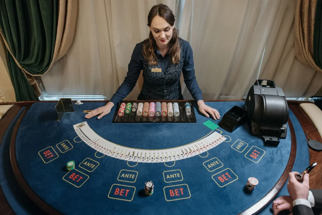 Croupier standing behind a blue baize gaming table with a fanned deck of cards and chip rack, in a room with heavy green and cream curtains