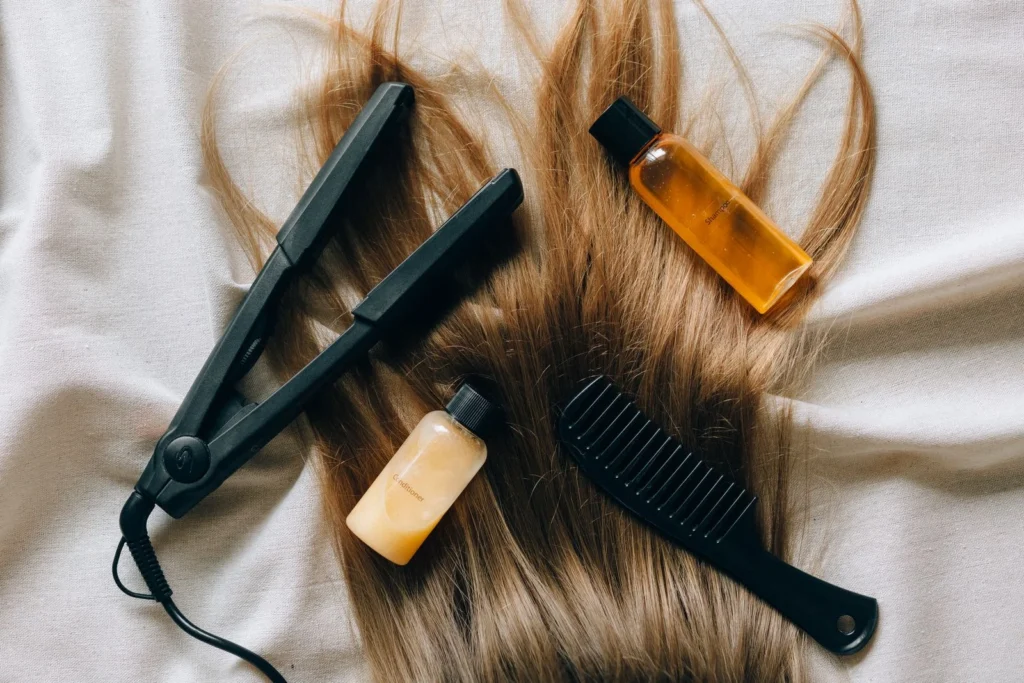 Why Good Haircare Starts With the Right Tools Black hair straightener, wide-tooth comb and bottles of hair oil laid over long light-brown hair