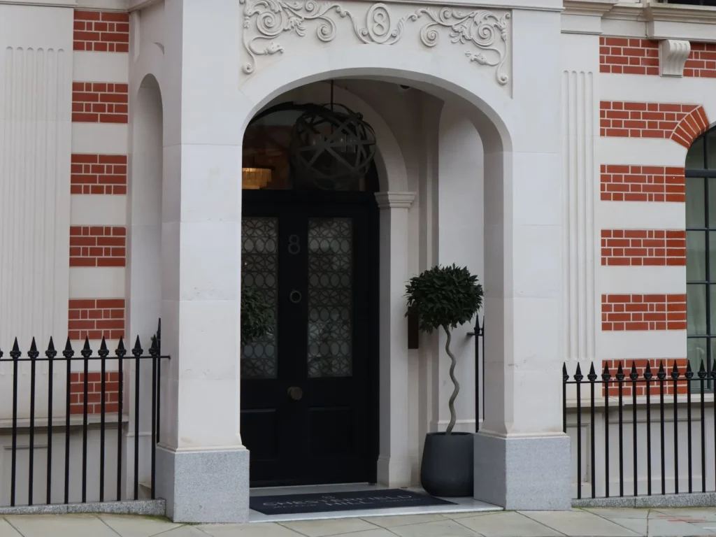 Arched stone doorway of a red-brick Mayfair townhouse with a black panelled door, topiary and iron railings